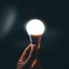 A close-up of a hand holding a bright LED light bulb against a dark background.